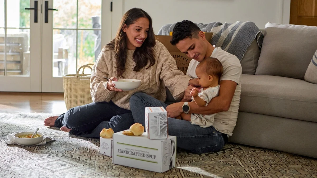 Parents sitting together on the floor with their baby, smiling and sharing a bowl of soup, with rolls and Spoonful of Comfort gift boxes nearby.