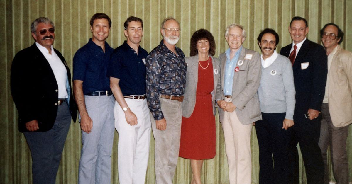 A line of men and one woman pose and smile for the camera