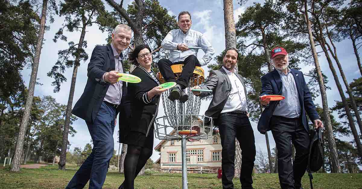 Four people in business casual clothing stand around a disc golf basket posing in throwing motions