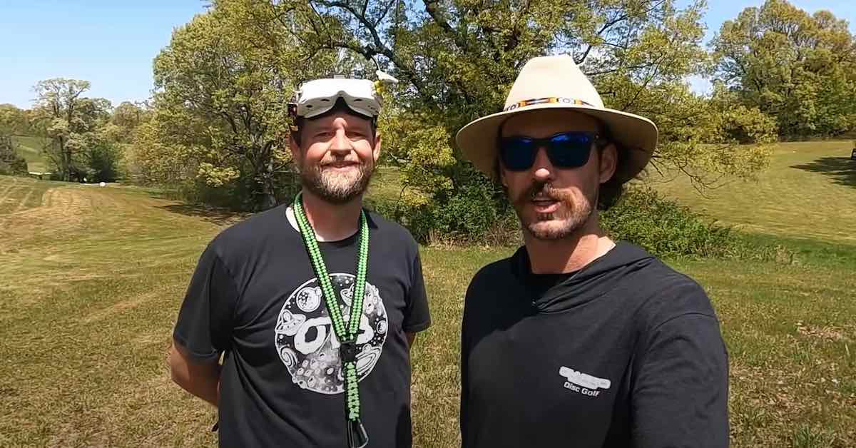 Two men look into a camera, one with a wide brim hat in the middle of speaking and the other smiling and wearing FPV goggles just over his eyes