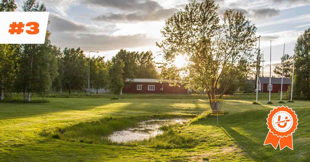 Gentle sunlight over a red wooden house at Skellefteå Discgolf Terminalen. Mowed green field. A yellow discgolf basket is under a small tree.