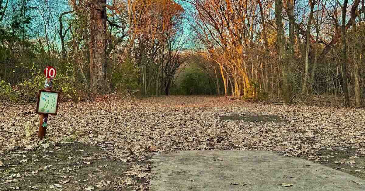 A concrete disc golf tee pad with rusted tee sign to the side and a tunnel fairway in the foreground