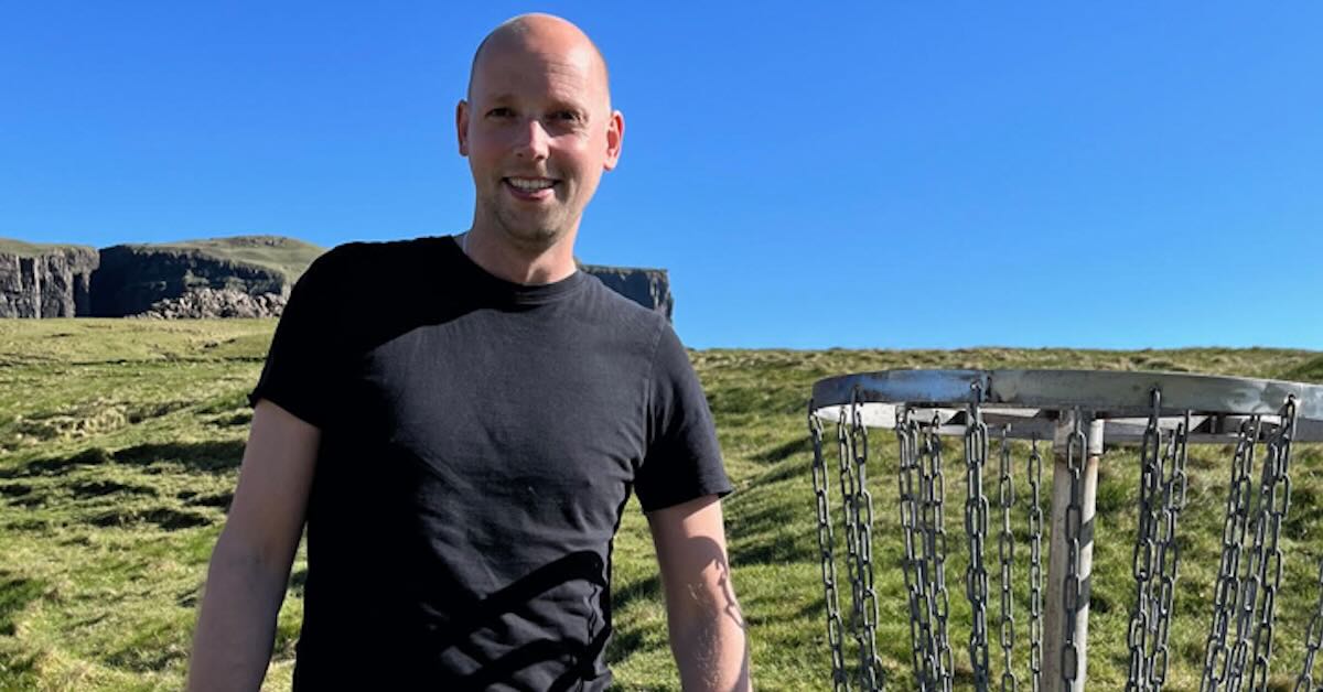 A man with a bald head and black t-shirt stands next to a disc golf basket in an open, grassy area
