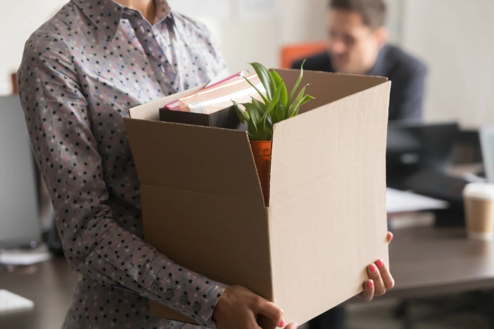 Woman in spotty shirt carrying a cardboard box of belongings from an office. A man works in the background