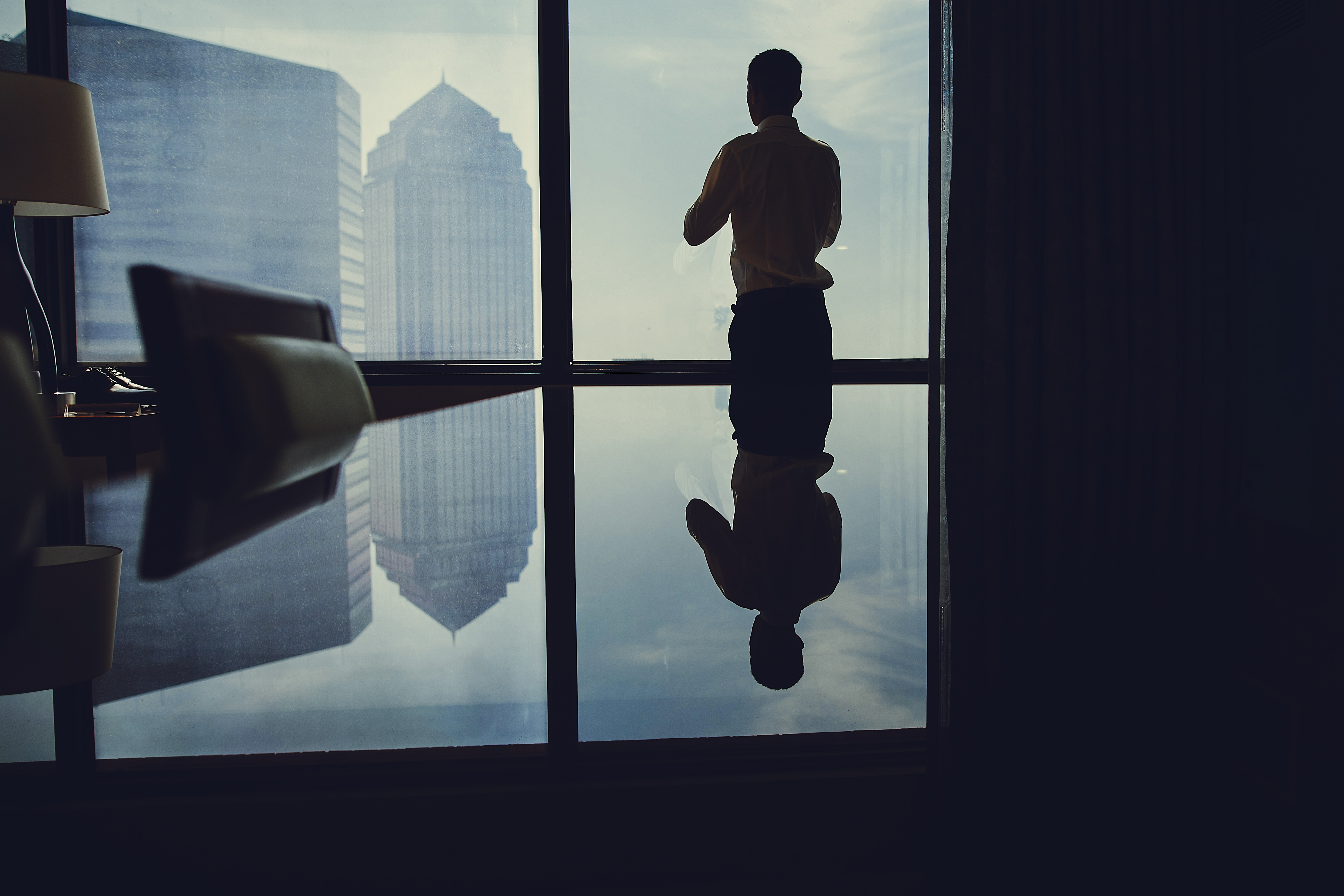 Businessman standing by a large office window, reflecting on a city skyline, contemplating business decisions.