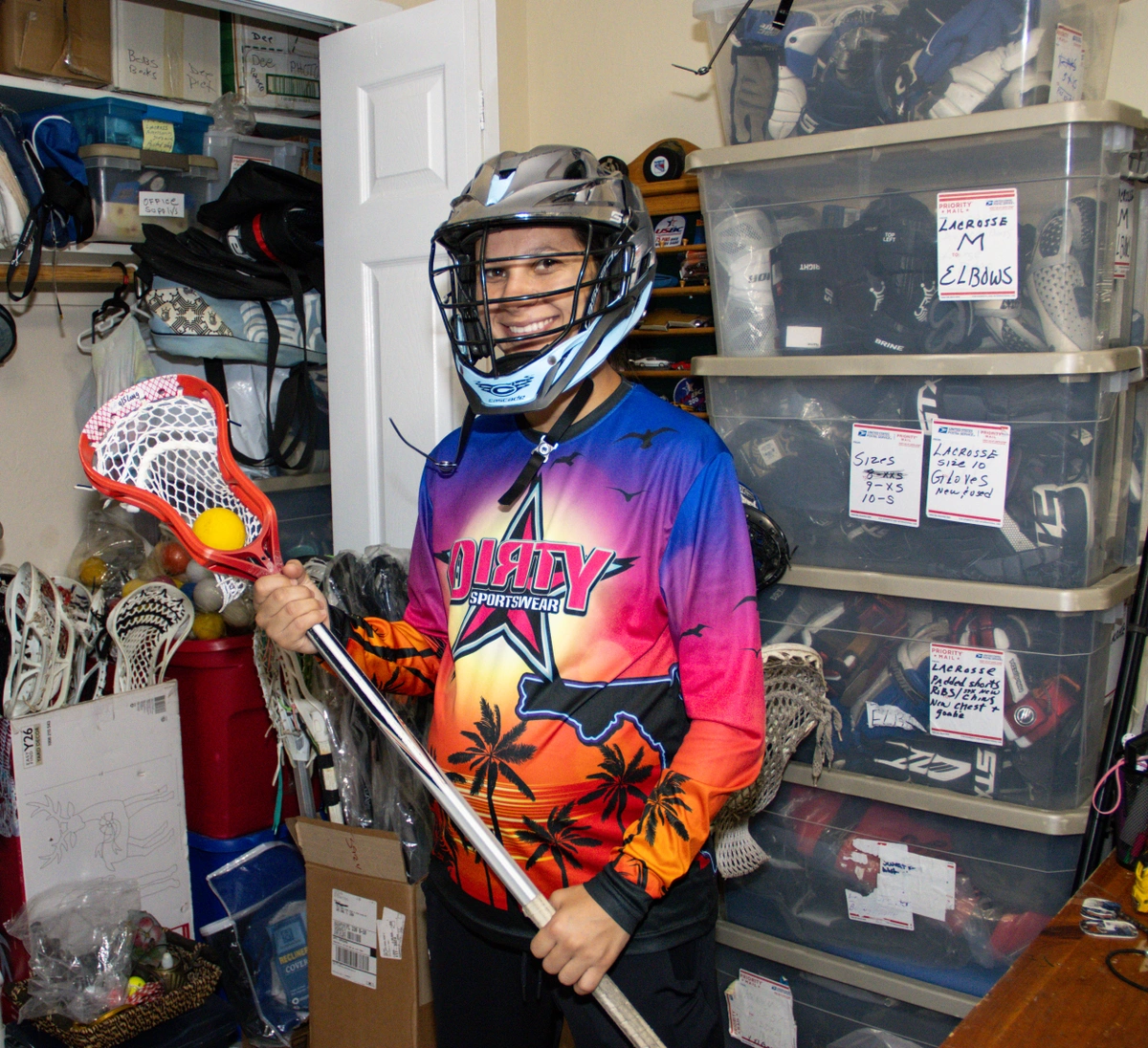 Tori Johnston holding a lacrosse stick wearing a lacrosse helmet in front of her lacrosse gear for sale