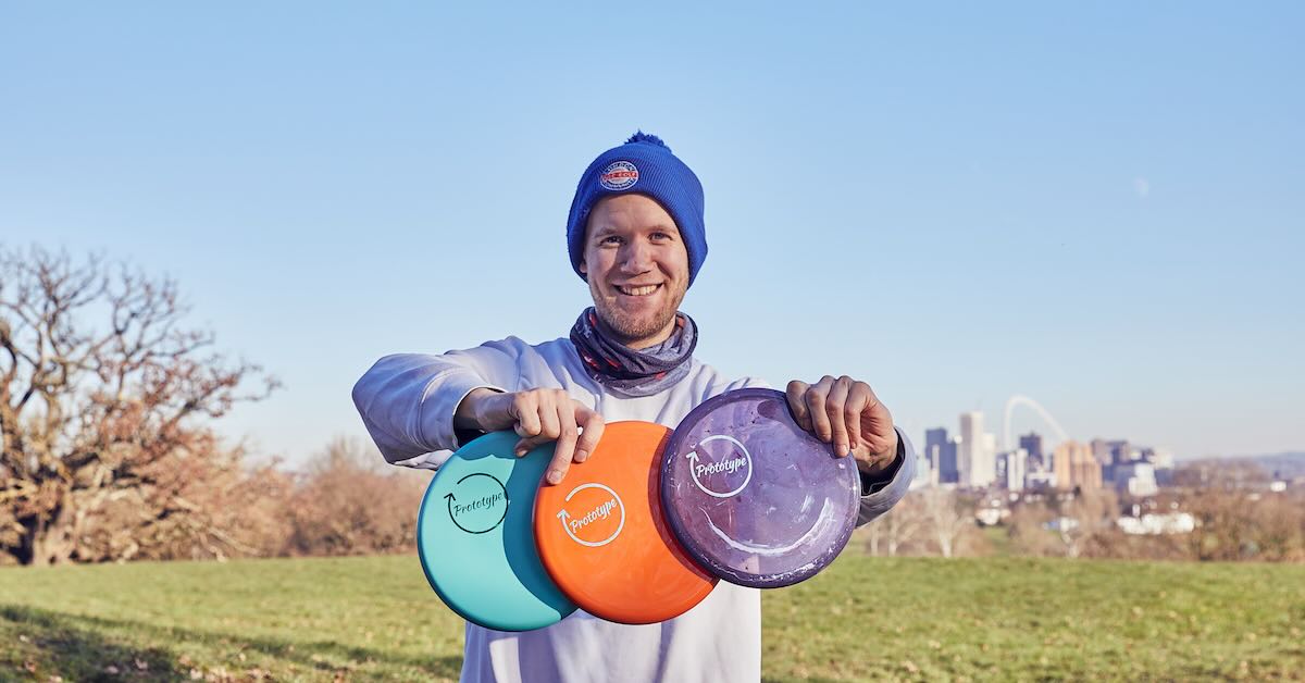 A man in a winter hat holds three discs in a park with a city skyline inthe backgorund