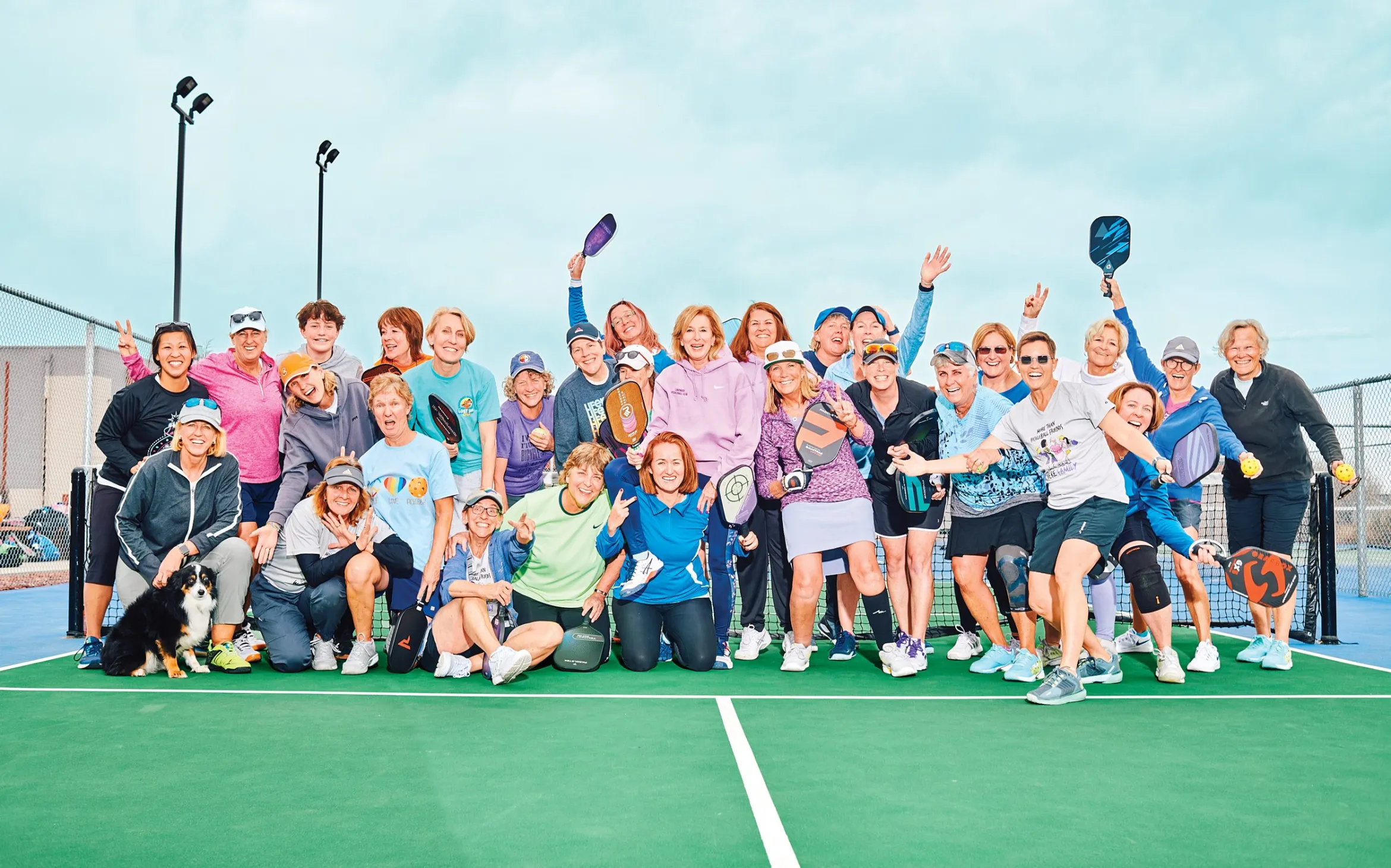 A group of women are smiling and holding pickleball paddles and balls on a pickleball court.