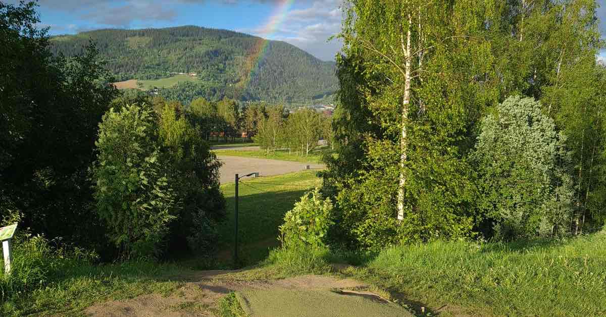 Turf disc golf tee pad leads to a large drop off with mountains and a rainbow in the distance