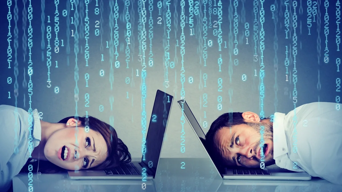 Two people rest their heads on open laptops as streams of glowing binary code fall across a blue-toned background, illustrating digital technology and cybersecurity.