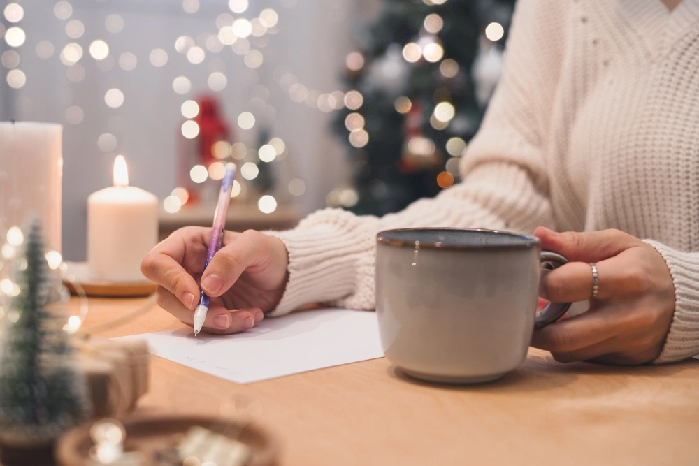 Woman in white sweater is writing a list while drinking from a mug. There's a Christmas tree, candles and twinkly fairy lights in the background.