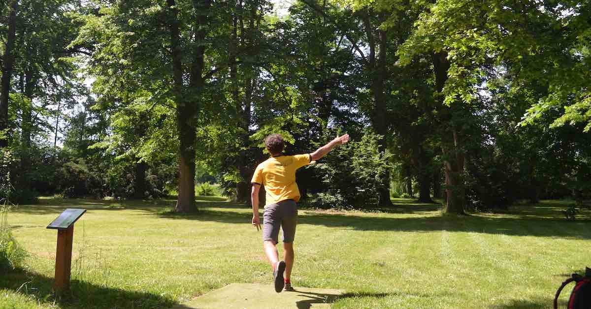 A player in a yellow shirt and shorts throws on a disc golf hole in a park setting with grass and many trees