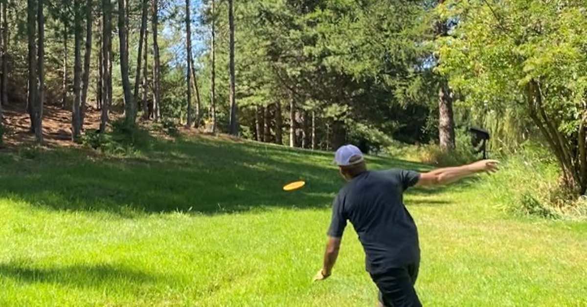 Man throwing a disc golf disc on a grassy fairway lined with trees