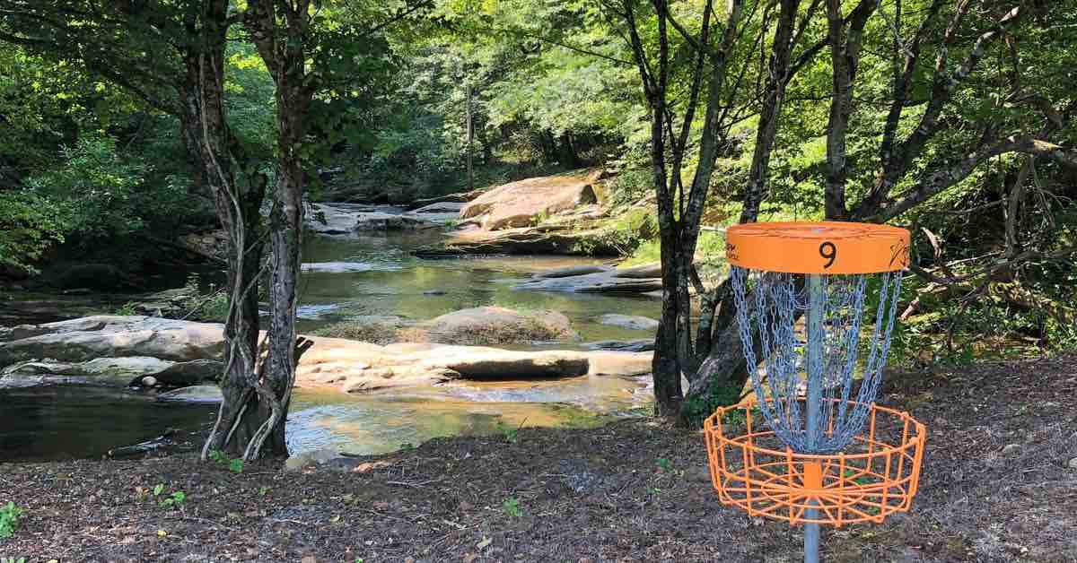 An orange disc golf basket in front of a rock-filled stream