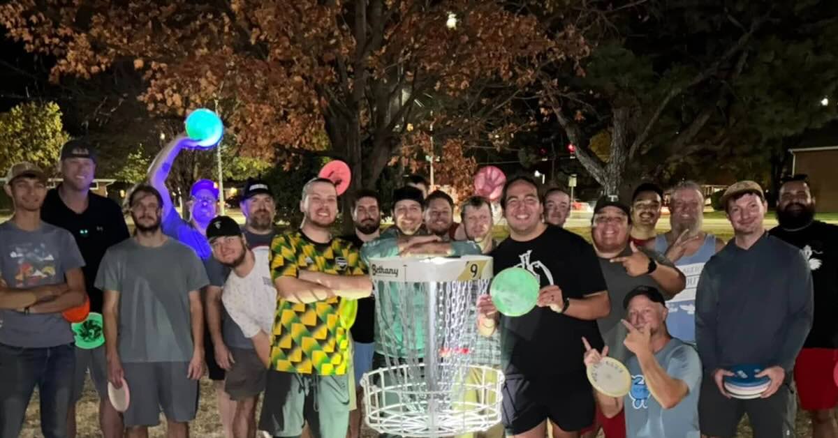 A group of disc golfers gathered around disc golf basket in a lit park at night