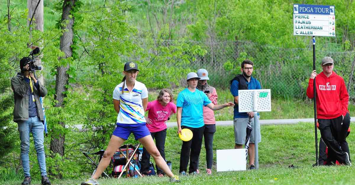 A woman stands on a disc golf tee pad as a small group looks on