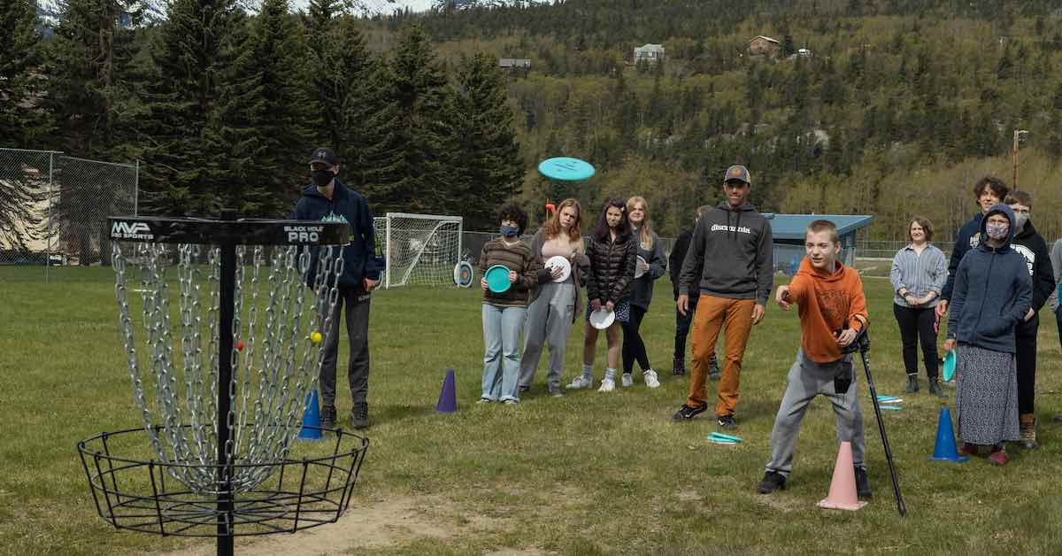 Young man on an athletic field putting at a disc golf basket with lines of other young people waiting behind