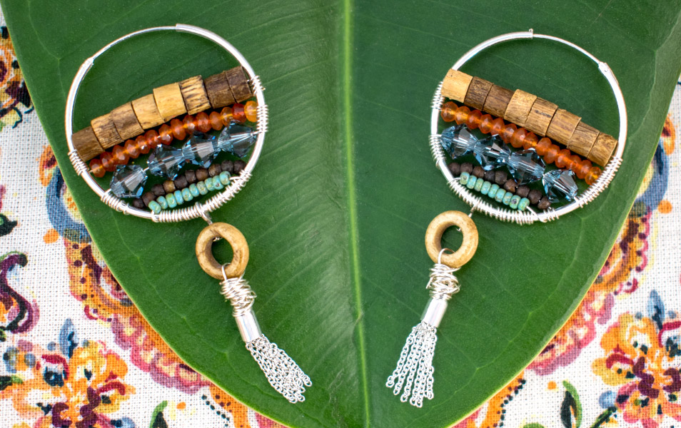 hoop earrings with multi colored beads wire wrapped across and a tassel sitting on a green leaf with flower fabric behind