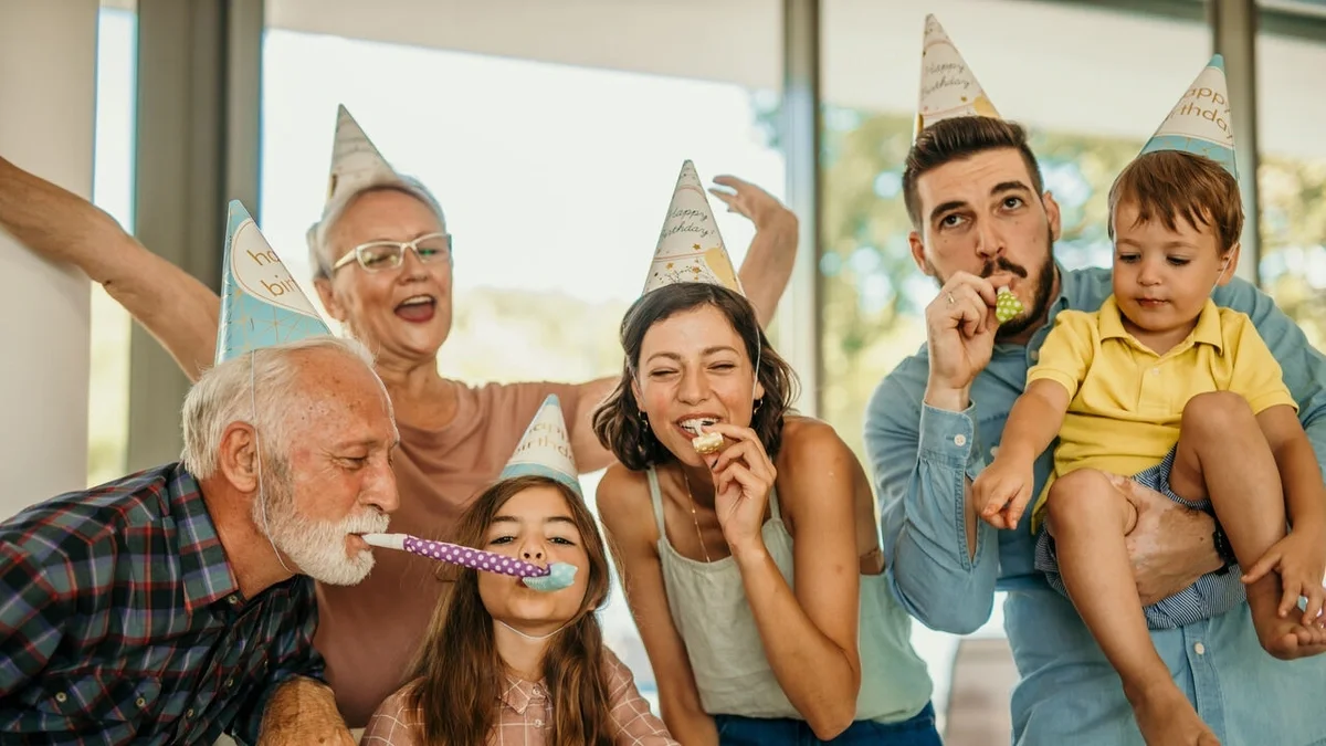 Multi-generational family wearing party hats, smiling and blowing party horns together while celebrating, with one adult holding a young child.