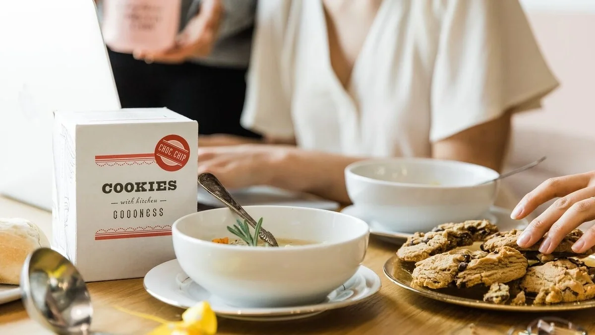 Close-up of a shared table with bowls of chicken soup, a plate of chocolate chip cookies, a box labeled “Cookies,” and people gathered together in conversation.