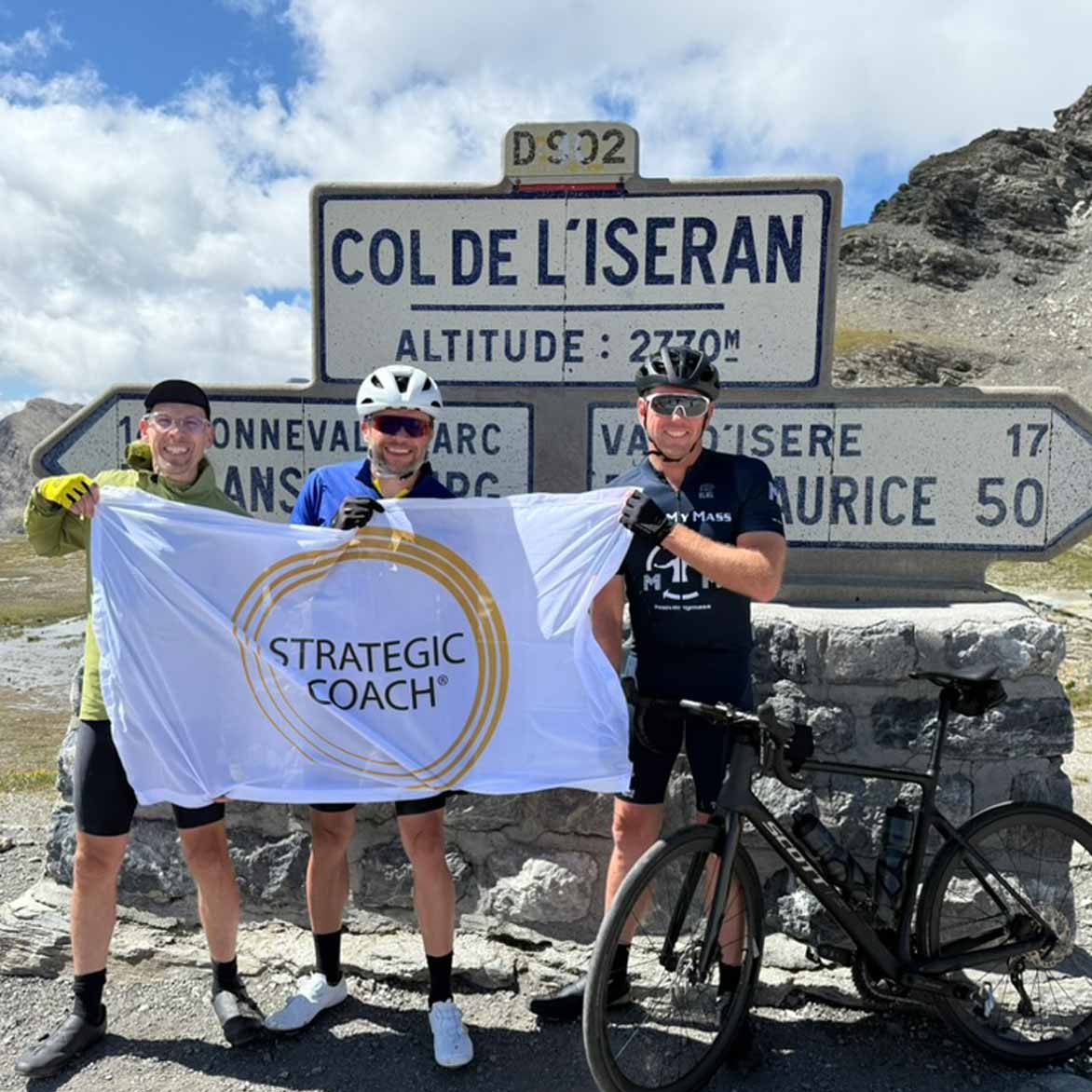 Strategic Coach member Kary Oberbrunner standing with friends at the top of Col De L’Iseran holding a Strategic Coach flag. 