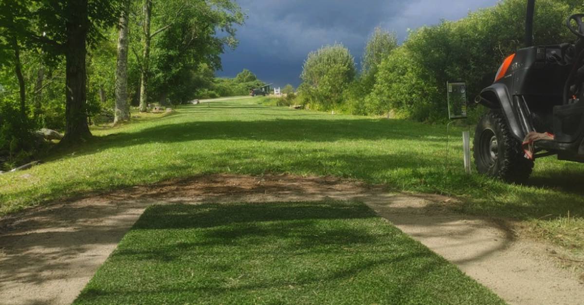 A turf disc golf tee pad in foreground a mixed bright but stormy sky in the background