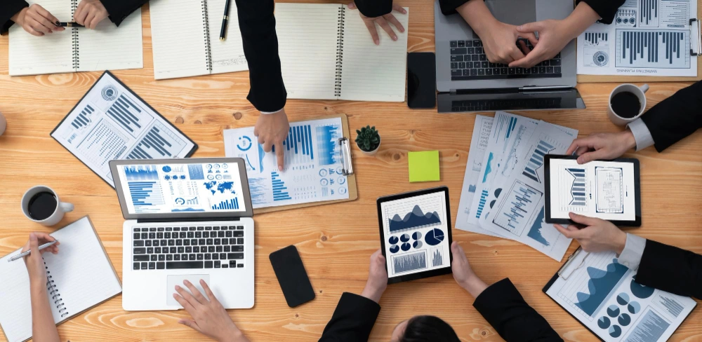 The image is a top-down view of a business meeting table with documents, digital devices, and hands of the attendees, engaged in analyzing charts and typing, accompanied by coffee cups and a small plant.