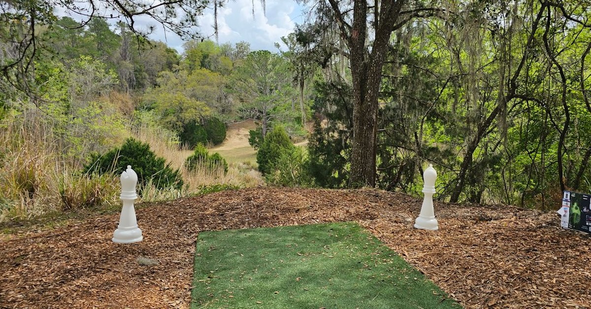 A turf disc golf tee at an elevated point overlooking a valley that rises sharply back up in the distance. Trees, grass all around.