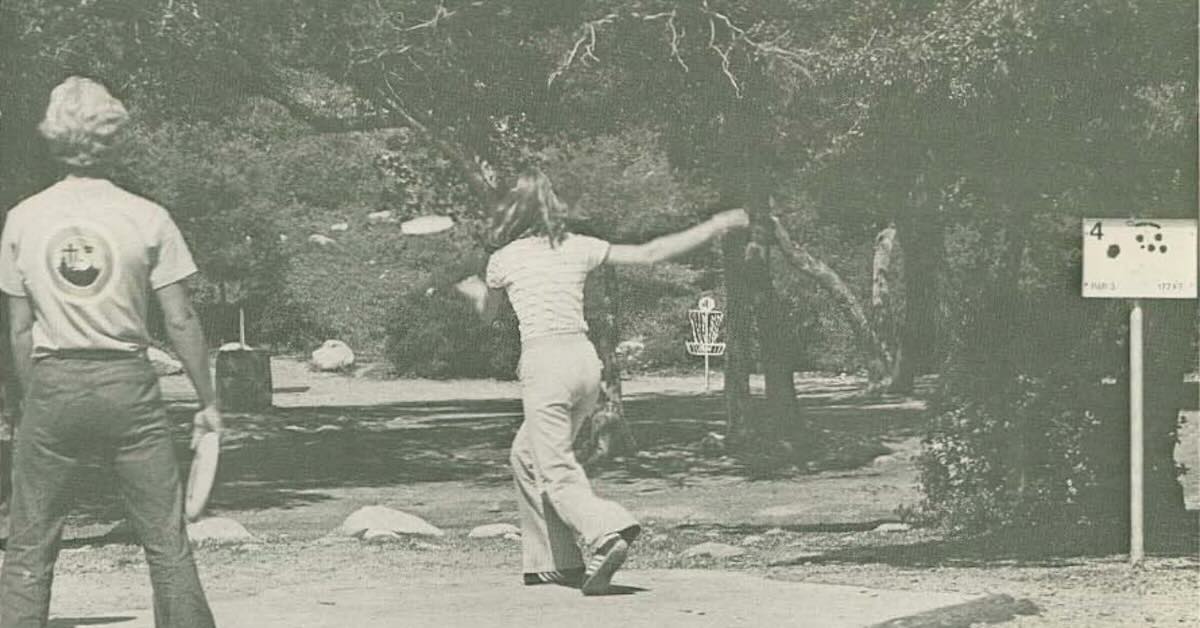 A black-and-white photo of a woman throwing a disc on an early disc golf hole with trees obstacles before a basket