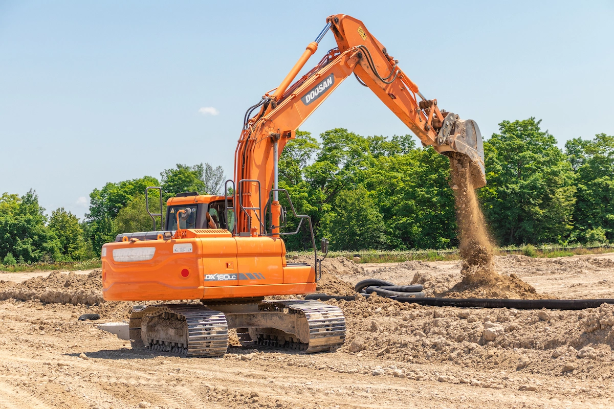 Orange Doosan excavator dumping dirt on a job site with trees in the background