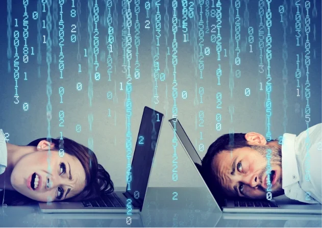 Two people rest their heads on open laptops as streams of glowing binary code fall across a blue-toned background, illustrating digital technology and cybersecurity.