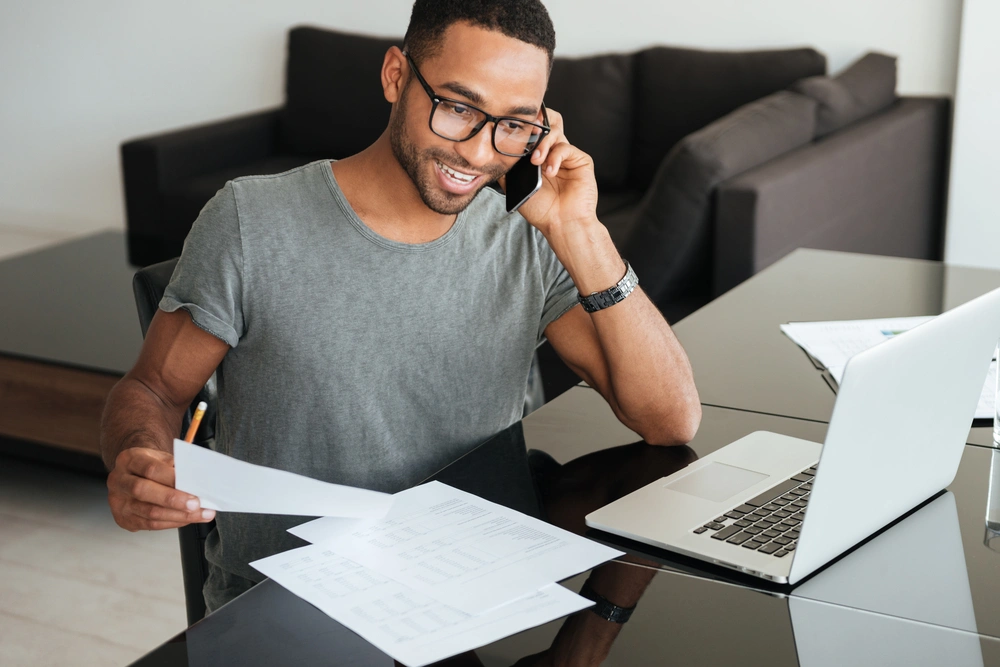 smiling man on the phone sat at a desk with his laptop and paperwork