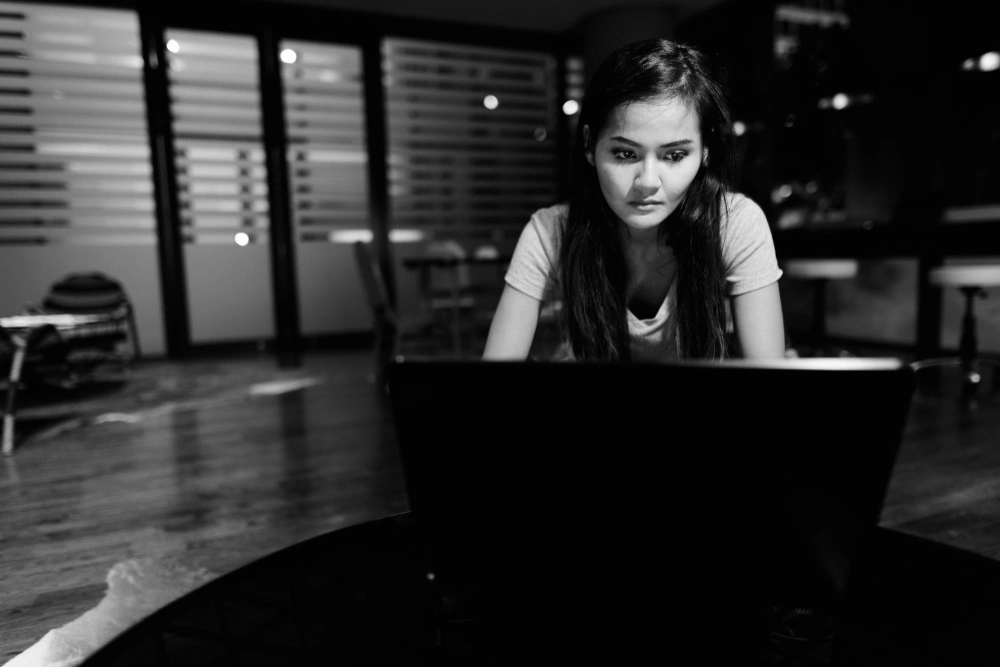 A woman focused on her laptop in a modern, dimly lit workspace.