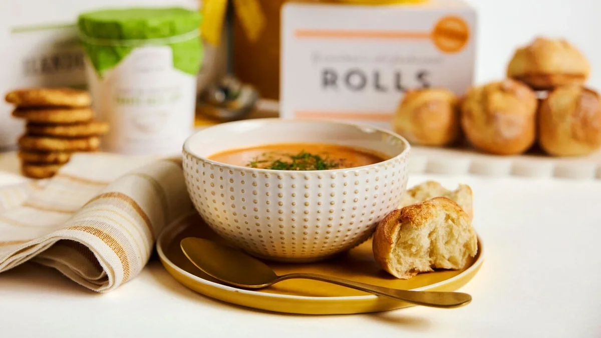 Close-up of a bowl of creamy tomato soup served with a fresh roll on a yellow plate, with rolls, cookies, and Spoonful of Comfort packaging softly blurred in the background.