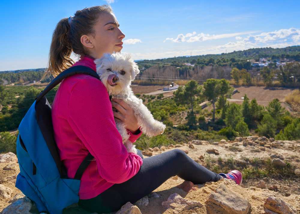 A woman in a pink jacket and blue backpack hugging a small white dog in nature