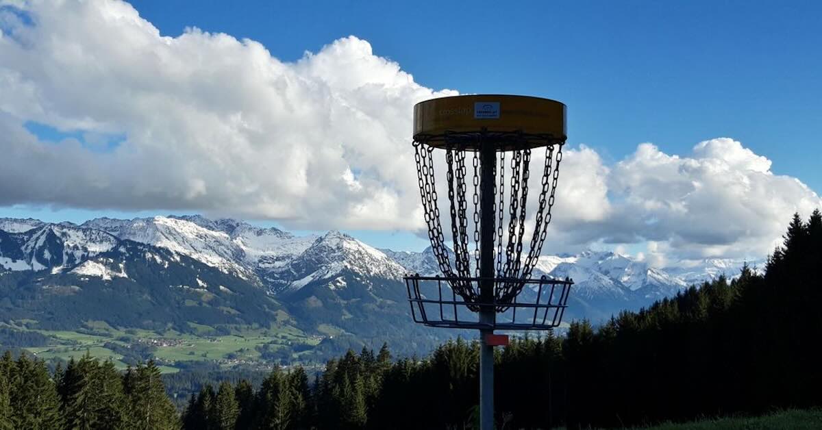 A disc golf basket with a stunning view of the Alps in clouds in the distance