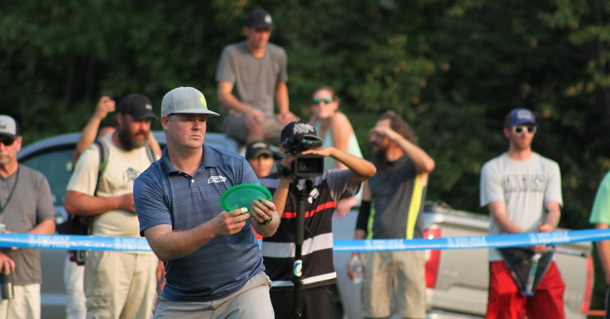 A crowd looks on as a man lines up a disc golf shot with disc in hand