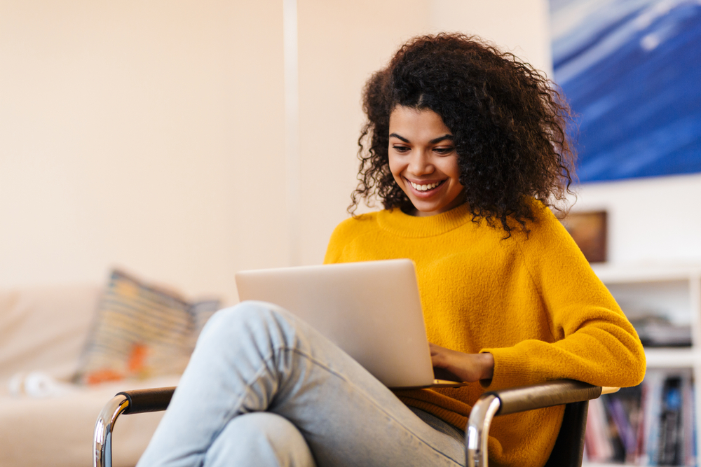Lady with curly hair in yellow jumper smiling while using a laptop