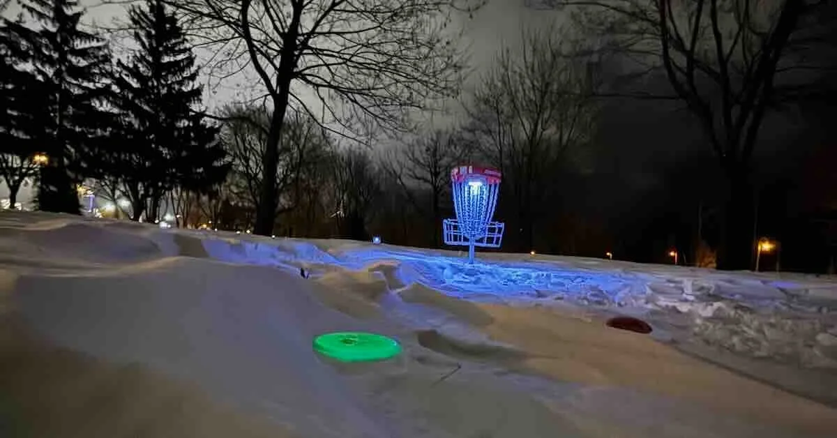 A disc glows green on thick snow as a disc golf basket is illuminated in the background at night in a park