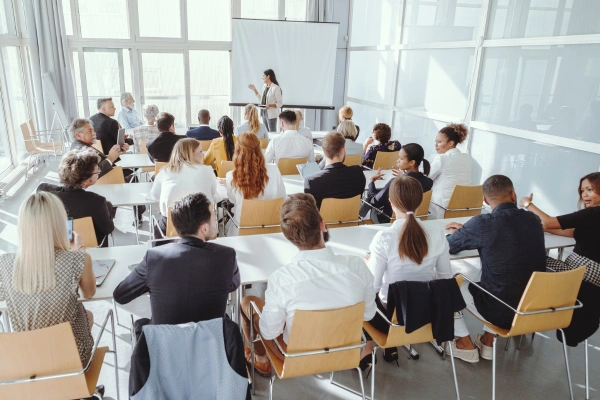 A room of workers sitting at long tables facing forward where a woman is presenting