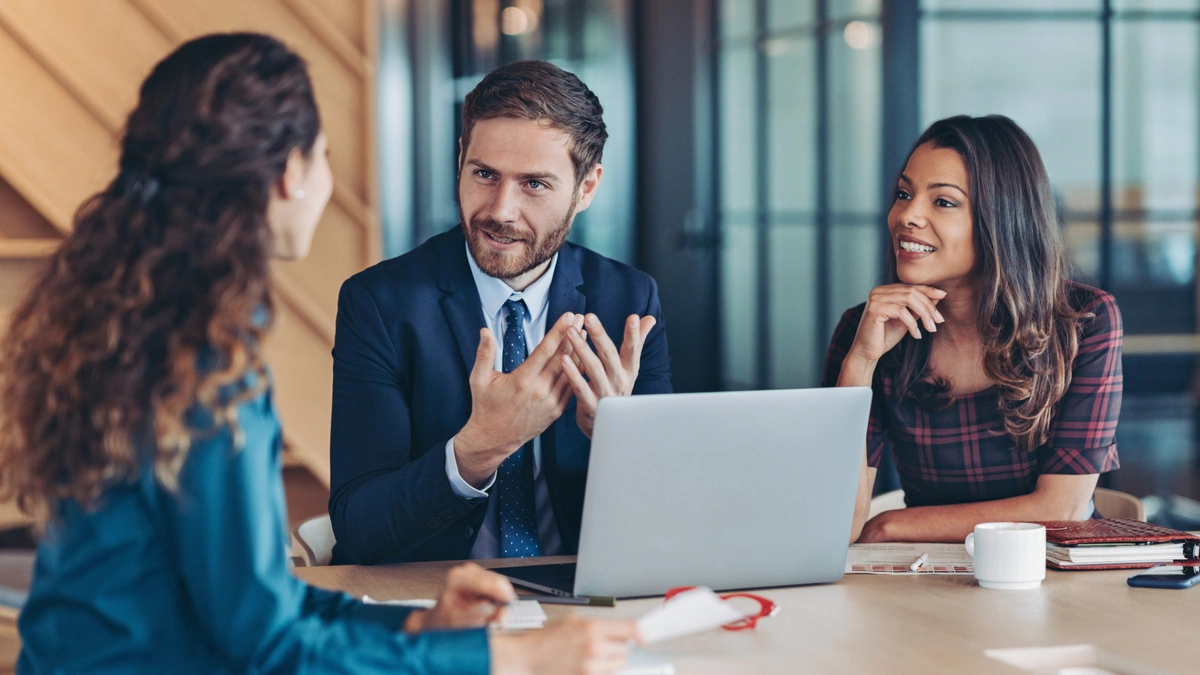 Three people sit at a table in an office, discussing. One gestures while speaking. An open laptop is on the table.