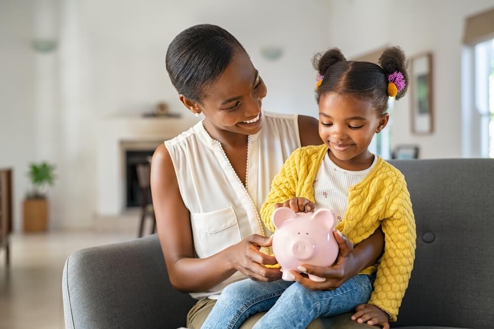 A black mother and daughter put money into a piggy bank and save money together.