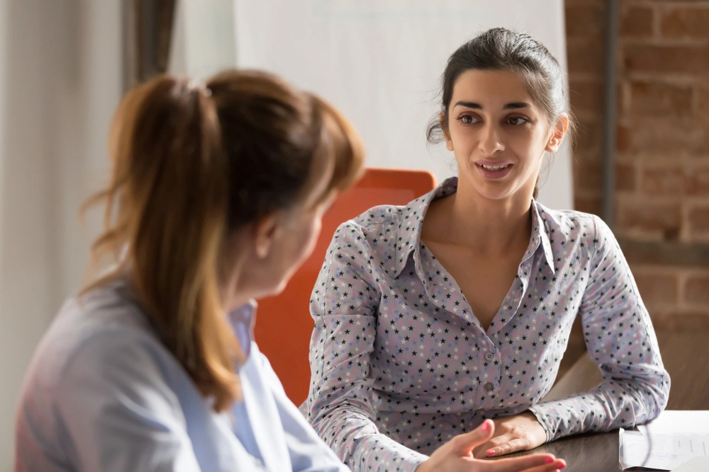 A dark haired lady in a spotted lilac shirt talks to a red haired lady with her back to the camera. They're both sitting at a desk.