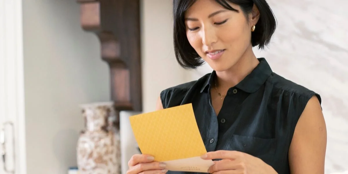 Woman smiling while reading a yellow greeting card indoors.