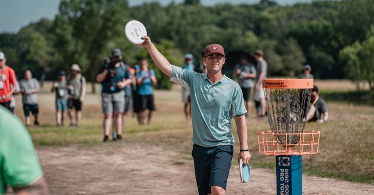 A man waves to the crowd at a disc golf event, putter in hand and near a disc golf basket