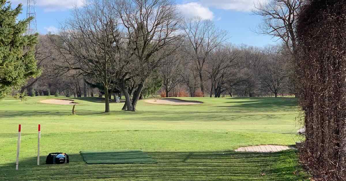 A turf disc golf tee pad on green grass on a ball golf course with bare, winter trees on a sunny day