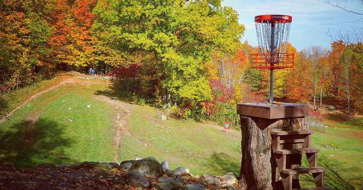 A red disc golf basket elevated on a tall stump in foreground with a background of a hill and trees in fall colors