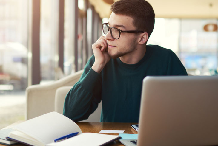Man in glasses looking out of the window with a notebook and a laptop on the table in front of him