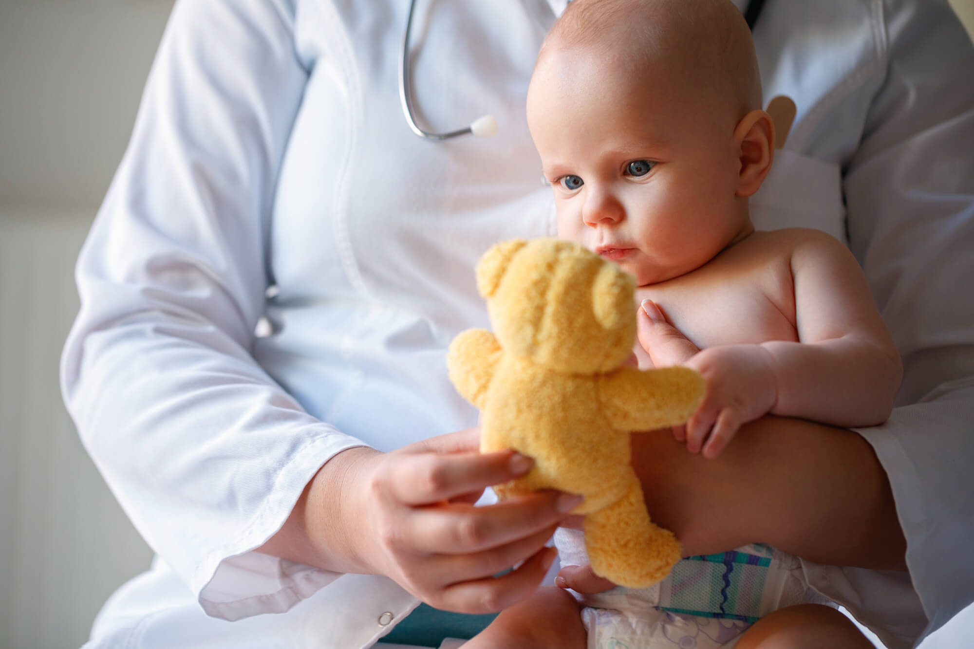 A little baby boy looking at a teddy bear while sitton on a pediatric doctor’s lap