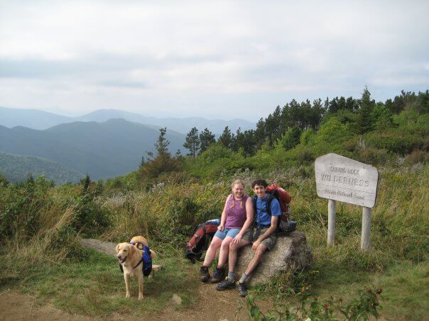 A Golden Retriever with a hiking backpack poses at a trailhead with a woman and a man in the mountains